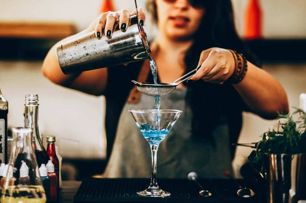 A bartender pouring a vibrant blue cocktail through a strainer into a glass.